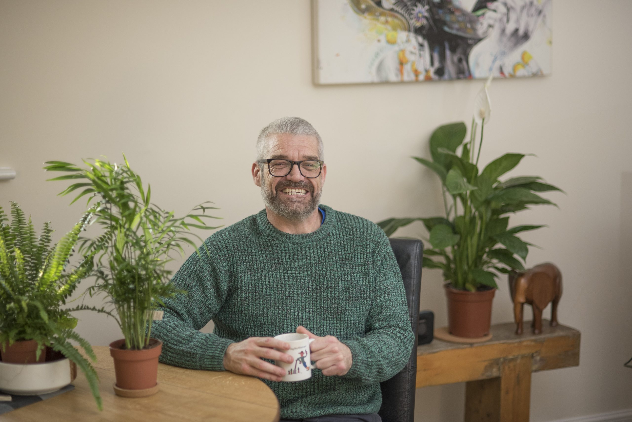 Travis sitting at his dining table, holding a mug, with plants in the background